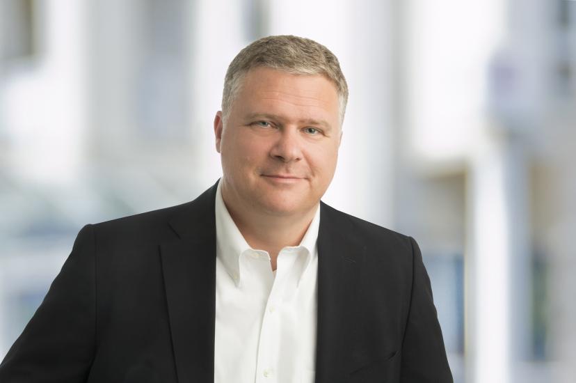  A professional headshot of a man with short, fair hair, wearing a black blazer over a white button-down shirt, set against a blurred indoor background.