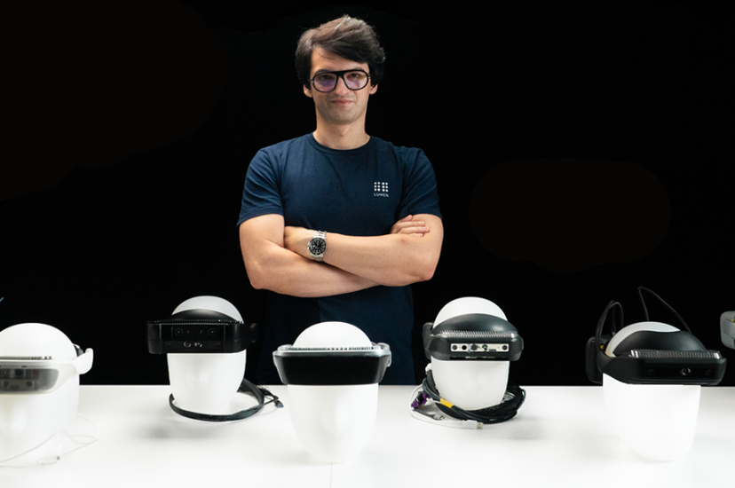 A white man with glasses and wearing a blue shirt stands behind a row of head mounted vision sesnors