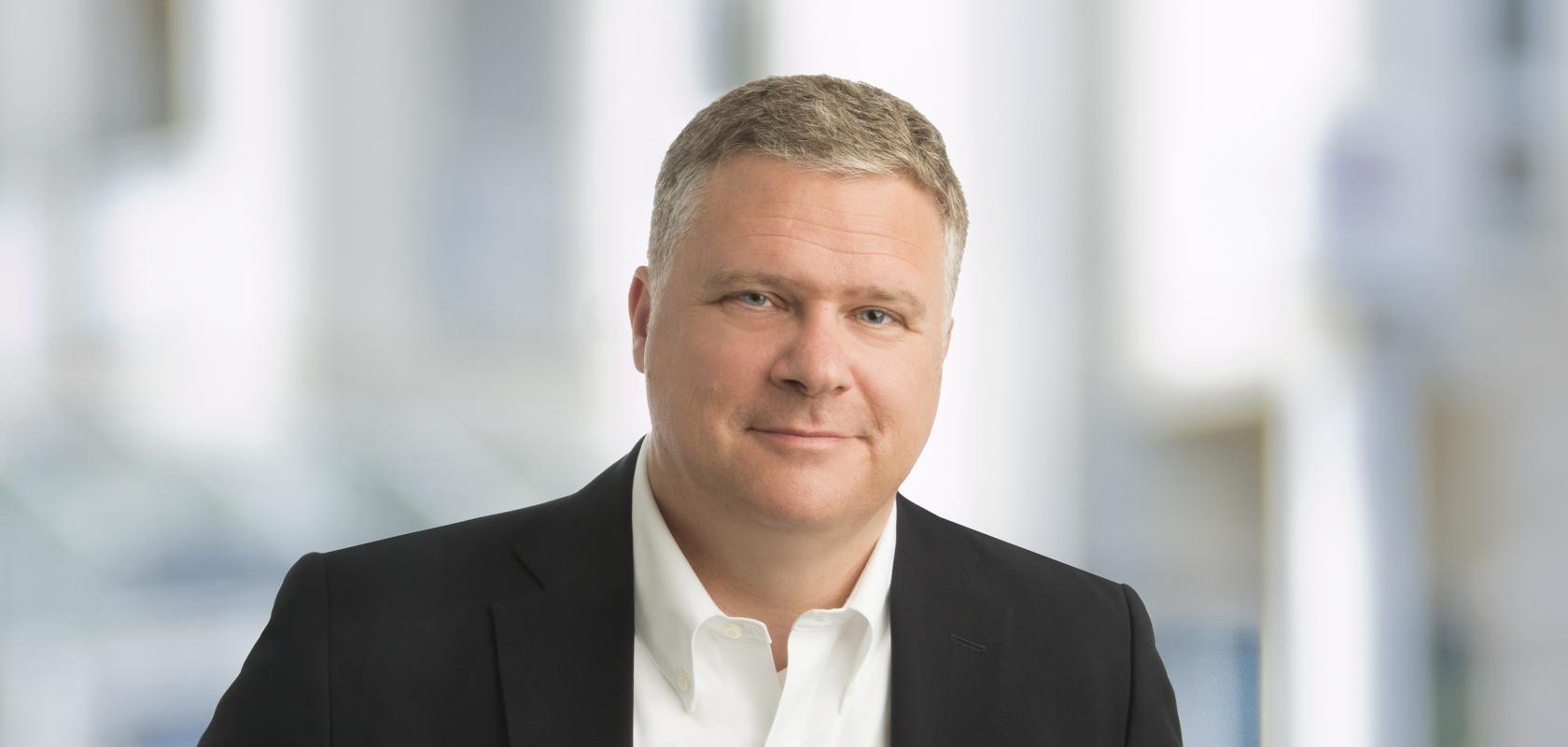  A professional headshot of a man with short, fair hair, wearing a black blazer over a white button-down shirt, set against a blurred indoor background.
