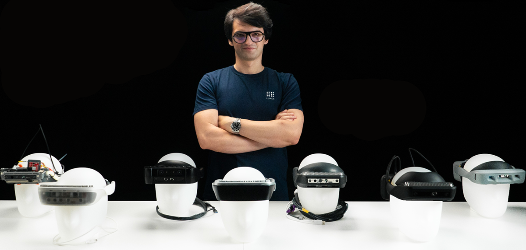 A white man with glasses and wearing a blue shirt stands behind a row of head mounted vision sesnors