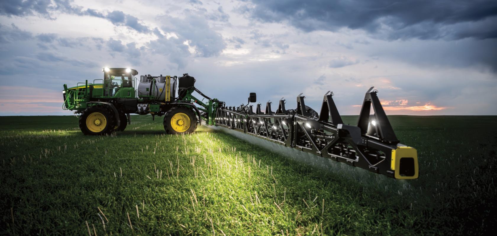 A green John Deere tractor spraying a field at dusk