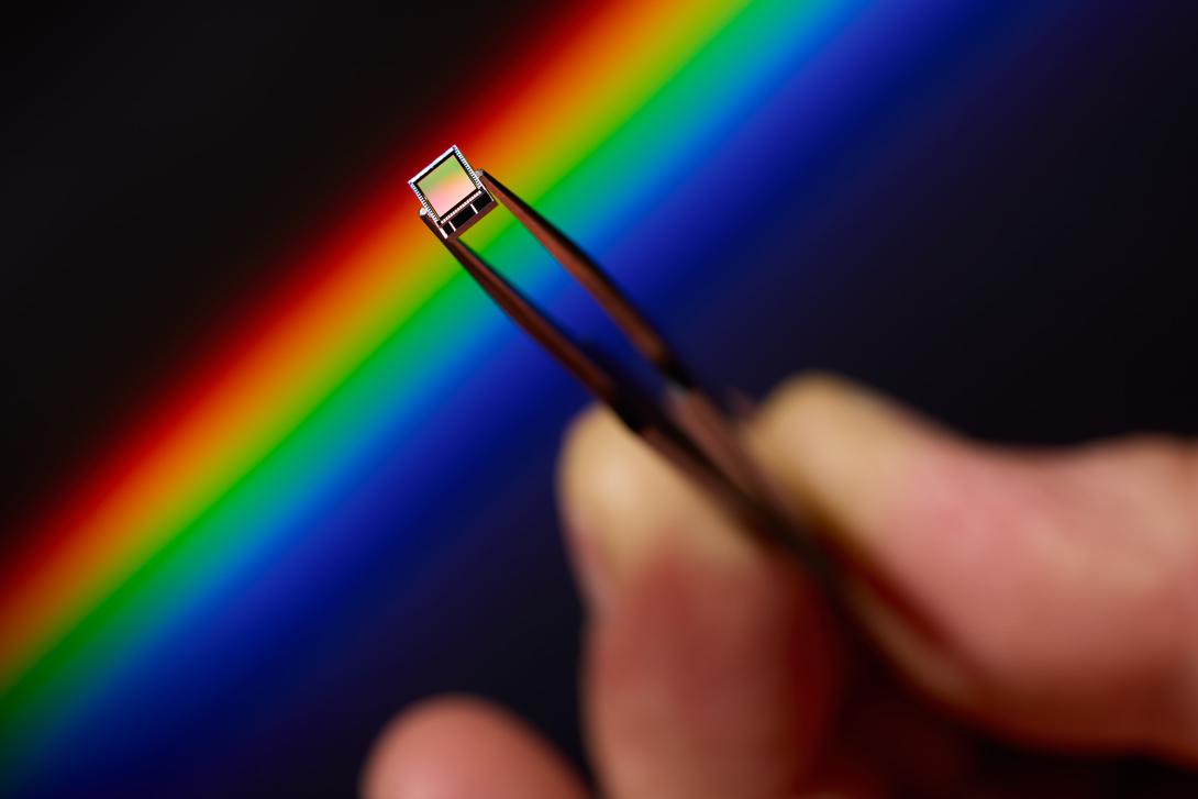 A hand holds a small image sensor in tweezers before a rainbow